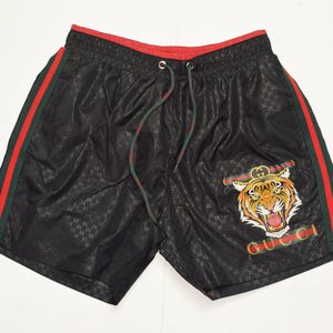 May include: Black athletic shorts with a red drawstring and red and green side stripes. The shorts feature a tiger graphic and the word "GUCCI" in red and green.