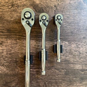 Może przedstawiać: Trzy chromowane klucze zapadkowe o różnych rozmiarach są prezentowane na tle drewnianym. Klucze, ułożone od największego do najmniejszego, mają polerowane metalowe wykończenie i czarne akcenty.