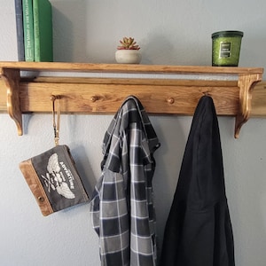 May include: A wooden shelf with a row of hooks, holding a small succulent in a white pot, two books, and a green candle. A gray and black plaid shirt and a black jacket hang from the hooks. A small pouch with the word "ADVENTURE" is also hanging.