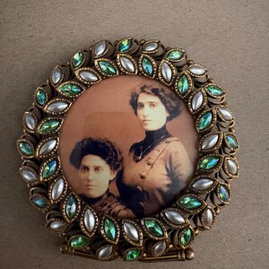 Puede incluir: Un marco de fotos redondo vintage con una fotografía en tonos sepia de dos mujeres. El marco dorado está adornado con joyas verdes e iridiscentes y detalles blancos perlados. El marco tiene un soporte para exhibición.