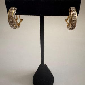 May include: A pair of gold-toned hoop earrings with a pave setting of small, clear stones. The earrings are displayed on a black velvet jewelry stand against a white background. The earrings have a hinged closure.