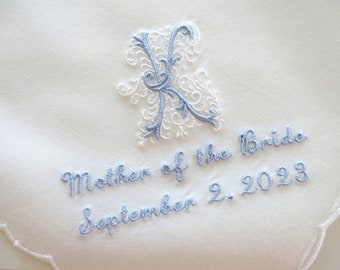 May include: A white fabric handkerchief featuring an embroidered light blue and white ornate letter K. Below the letter, the text "Mother of the Bride" and "September 2, 2023" is embroidered in light blue thread. The fabric has a scalloped edge.