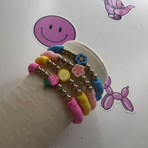 May include: A stack of colorful beaded bracelets with charms, including a strawberry, lemon, flower, and a blue flower. The bracelets are on a white surface.