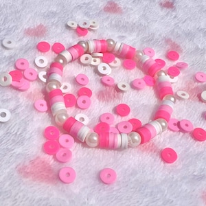 Puede incluir: Un brazalete de cuentas rosa y blanco con detalles de perlas blancas. El brazalete está rodeado de cuentas rosadas y blancas en forma de rosquilla.