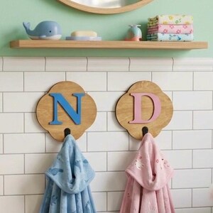 May include: Two wooden wall hooks shaped like four-leaf clovers, each featuring a large letter, N in blue and D in pink. A blue hooded towel with a pattern hangs from the N hook, and a pink hooded towel with a pattern hangs from the D hook.