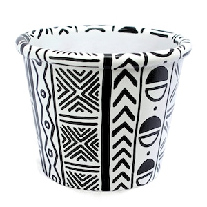 May include: A white ceramic planter with a black geometric pattern. The design includes zigzags, chevrons, and half-circle shapes. The planter has a rounded top and a cylindrical shape, suitable for indoor or outdoor use.