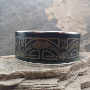 May include: A black cuff bracelet with a silver trim and a decorative floral and geometric pattern. The bracelet is resting on a gray stone surface. The design features symmetrical floral elements.
