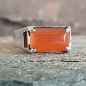May include: A silver ring featuring a rectangular, smooth, orange carnelian gemstone set in a four-prong setting. The band is polished silver, and the stone is positioned horizontally, showcasing a vibrant, translucent orange hue.