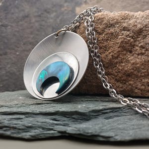 以下が含まれることがあります： シルバーのペンダントネックレス。ペンダントは、大きな楕円形、小さな楕円形、虹色の青と緑の色合いの三日月型の象嵌を備えた、レイヤードデザインです。チェーンもシルバーです。