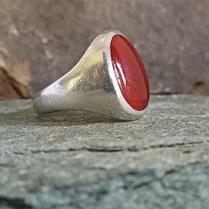 May include: A silver signet ring featuring a smooth, oval-shaped, vibrant red stone set into the center. The metal band has a polished finish with visible surface texture and light scratches, showcasing a classic, solid construction.