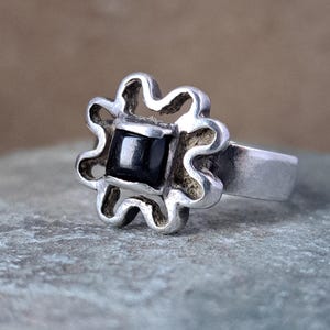 May include: A silver ring with a flower-shaped setting. The setting has a square, black stone in the center. The ring's band is a simple, smooth design.