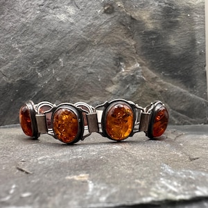 Könnte beinhalten: Ein silbernes Armband mit ovalen Bernsteinen. Die Bernsteine haben eine warme, durchscheinende orange-braune Farbe und sind in einen silbernen Metallrahmen gefasst. Das Armband wird auf einer dunkelgrauen, strukturierten Oberfläche präsentiert.