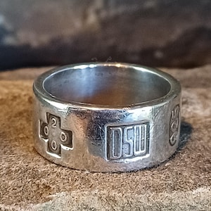 May include: A silver band ring with a textured finish. The exterior features a stamped cross symbol containing the number 2000, a rectangular box with the letters DSW inside, and the number 925 stamped on the side of the metal band surface.