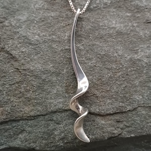 May include: A silver pendant necklace featuring a swirling, spiral design. The pendant has a polished, curved form that twists and narrows. The necklace chain is a fine, linked style. The jewellery is shown against a grey stone backdrop.