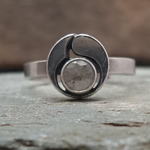 May include: A silver ring with a unique design. The ring features a circular setting with a central, round, light-colored stone. The setting has an abstract, curved design. The band is a simple, polished silver. The ring is displayed on a textured surface.