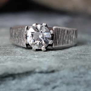 May include: A silver-toned ring featuring a single round, clear faceted gemstone set in a six-prong mounting. The band has a distinct vertical textured finish on both sides of the central stone, providing a contrast to the smooth metal prongs.