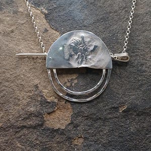 May include: A silver pendant necklace featuring a half-circle design with a textured floral pattern and two concentric hammered rings. The pendant is suspended from a delicate chain, creating a modern and artistic jewelry piece.