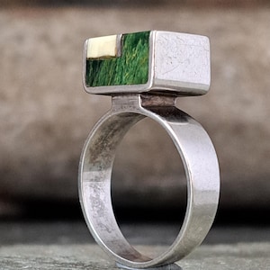 May include: A silver ring featuring a rectangular top section inlaid with a textured green panel and a smaller cream-coloured rectangular accent. The band is smooth and polished, with a wide, flat profile that supports the prominent rectangular setting.