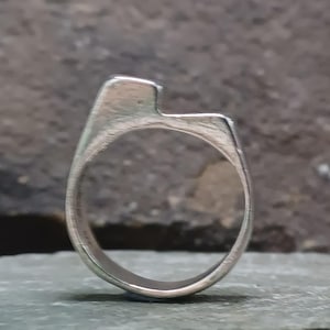 May include: A silver-toned metal ring with a brushed finish, featuring an asymmetrical top section with a raised rectangular element on one side and a flat, horizontal extension on the other, creating a distinct open-top silhouette.