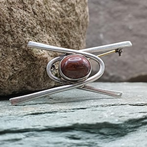 May include: A silver-toned brooch with a central, round, reddish-brown stone. The brooch has a geometric design with intersecting bars and a looped element around the stone. The pin is set against a textured, gray-green stone surface.