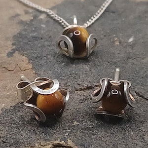 May include: A jewelry set featuring a necklace pendant and two stud earrings. Each piece has a silver-colored metal setting that cradles a round, polished, brown gemstone. The necklace has a delicate chain.