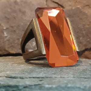 May include: A ring featuring a large, rectangular, faceted orange gemstone set in a dark, metallic band. The stone has a polished, reflective surface with sharp edges, while the band has a matte, weathered finish with a triangular side profile.