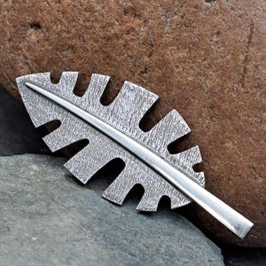 May include: A silver-toned metal brooch shaped like a stylized leaf with a textured, matte surface and a smooth, polished central vein running the length of the piece. The design features symmetrical, rectangular cutouts along both sides.