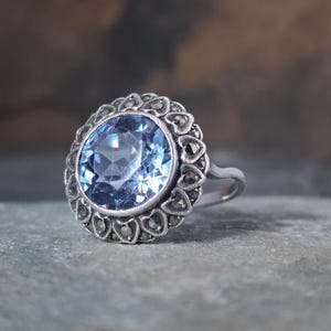 May include: A vintage silver ring featuring a large, round, light blue gemstone. The gemstone is surrounded by a halo of smaller, heart-shaped accents. The ring has a classic, elegant design.