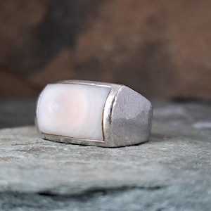 May include: A silver ring with a rectangular, off-white stone. The stone has a subtle pink hue in the center and is framed by a thin gold border. The ring's band has a textured, hammered appearance.