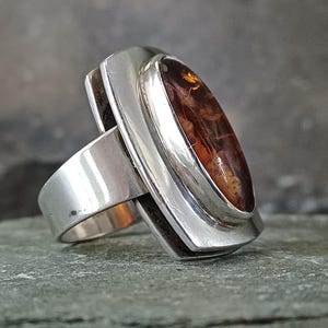 May include: A silver ring featuring a large, oval-shaped, polished amber gemstone set in a smooth metal bezel. The ring has a wide, solid silver band that supports the elevated setting, showcasing the deep orange and brown hues of the amber.