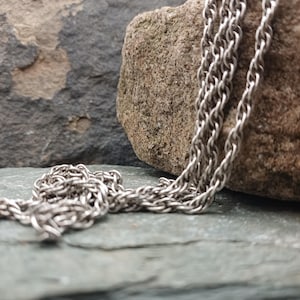 Op de afbeelding: Een zilverkleurige ketting met een geweven ontwerp. De ketting is gedrapeerd over een ruwe, bruinachtige steen, tegen een achtergrond van grijze en groene steen. De schakels van de ketting zijn duidelijk zichtbaar.