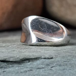 May include: A polished silver ring with a smooth, undulating surface and a wide, asymmetrical band. The metal reflects light across its curved, organic shape, showcasing a continuous, solid construction with no visible stones or engravings.