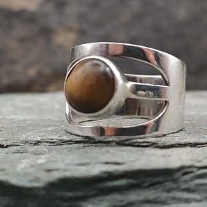 以下が含まれることがあります： 茶色のタイガーアイの石が付いたシルバーリング。リングは、側面に切り欠きのあるモダンなデザインです。楕円形の石はベゼルセッティングでセットされています。バンドは幅が広く、滑らかです。