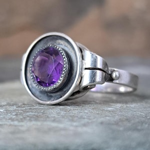 May include: A silver ring featuring a large, faceted, round, purple gemstone. The gemstone is set within a circular bezel, surrounded by a beaded border. The ring has a unique, hinged design.