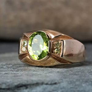 May include: A gold ring with a large, oval-shaped peridot gemstone at its centre. The ring has a classic design with smaller peridot accents on the sides, giving it a vintage appearance. The ring's band is of a substantial width.