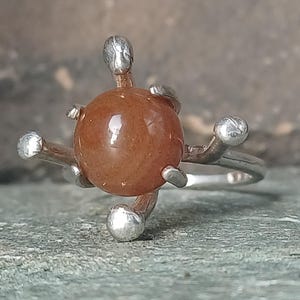 Könnte beinhalten: Ein Silberring mit einem zentralen, glatten, runden, rotbraunen Stein. Der Stein wird von vier silbernen Krappen gehalten, die nach außen ragen und jeweils in einer kleinen, abgerundeten, metallischen Kugel enden.