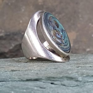 May include: A silver ring featuring a large, oval-shaped abalone shell inlay with iridescent blue, green, and purple tones. The band has a wide, open-work design that curves upwards to hold the polished shell setting securely in place.