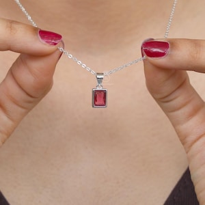 Puede incluir: Un collar de plata con un colgante rectangular de piedra roja. El collar lo sostiene una persona que lleva una camiseta marrón oscuro. Las uñas de la persona están pintadas de rojo.