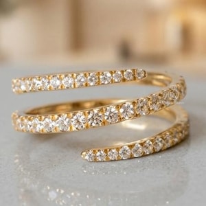 Puede incluir: Un anillo de oro con un diseño en espiral que se envuelve alrededor del dedo. La banda está incrustada con numerosas piedras pequeñas, claras y de corte redondo que cubren toda la superficie, creando un efecto brillante continuo.