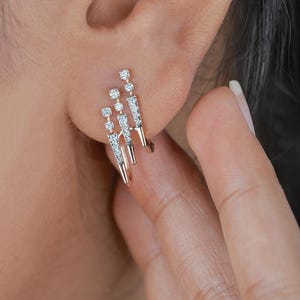 May include: Close-up of a pair of earrings with a unique design. Each earring features three vertical bars, each adorned with small, sparkling stones. The earrings are gold-toned, and the stones appear to be clear, creating a sophisticated look. The earrings are worn on an ear.