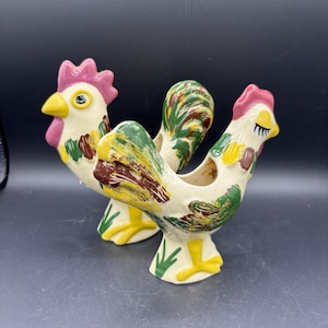 May include: Two ceramic rooster figurines. The roosters have a cream-colored base with colorful accents of green, yellow, brown, and pink. The roosters have yellow feet, pink combs, and yellow beaks. The figurines are on a dark background.