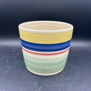 May include: A ceramic planter with a cream-colored interior and a colorful exterior. The exterior features horizontal stripes in yellow, blue, red, and green. The planter is cylindrical and has a smooth finish.