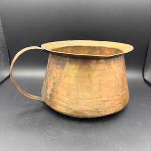 May include: A copper-colored metal mug with a handle. The mug has a wide, slightly flared rim and a rounded body. The metal has a mottled appearance with areas of darker and lighter tones, suggesting age or patina. The handle is curved and attached to the side of the mug.
