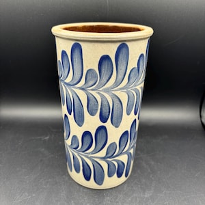 May include: A tall, cylindrical ceramic vase with a cream-colored base and a brown interior. The exterior is decorated with a repeating pattern of blue, leaf-like designs. The vase has a simple, rounded rim.