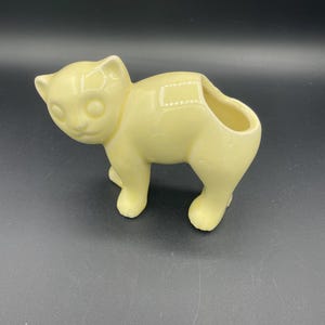 May include: A pale yellow ceramic cat planter with a hole in the back for drainage. The cat is standing on all fours with its head tilted slightly to the side.