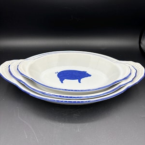 May include: Set of three oval serving dishes. The dishes are white with a blue pig silhouette in the center. Each dish has a blue rim and a decorative edge. The dishes are stacked on top of each other.