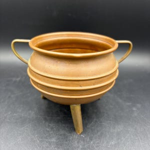 May include: A small, antique copper cauldron with a rounded body and three short legs. The cauldron has two small handles and horizontal bands around its midsection. The copper has a warm, aged patina, suggesting it is vintage.