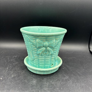 May include: A turquoise ceramic planter with a basket weave design and a bow detail. The planter includes a matching saucer. The planter is approximately 5 inches in diameter and 4 inches tall.