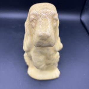 May include: A ceramic dog figurine in a light yellow color. The dog has long ears and a detailed face. The figurine has a crackled glaze finish, giving it a vintage appearance. The dog is sitting upright.