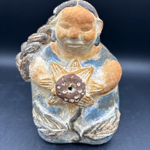 May include: A ceramic figurine of a seated figure holding a sunflower. The figure has a light brown face and hair, with blue and tan clothing. The sunflower is tan and brown. The figurine is on a dark background.